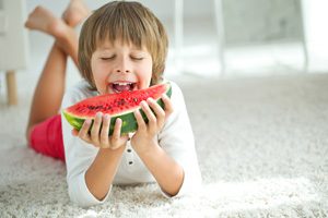 Kind liegt auf hellem Teppich und beißt in eine Melone